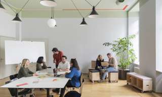 Blick ins Atelier im Berner Generationenhaus. Mehrere Menschen sitzen an einem weissen Tisch oder auf Holzelementen und diskutieren.